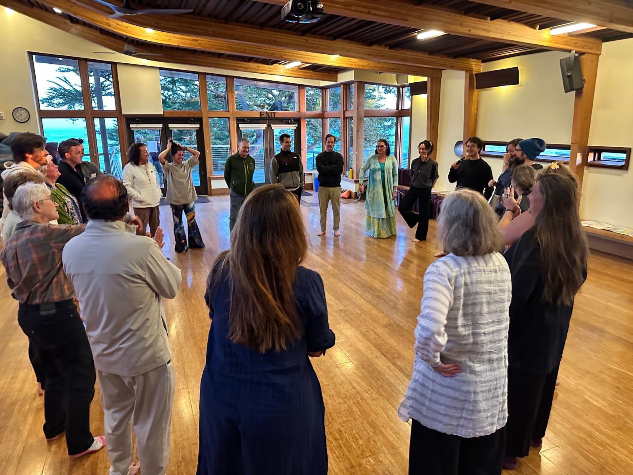 Liam holding a microphone, leading improv games for a large standing circle at a breathwork event at Esalen, in a wood-floored hall with ocean views.
