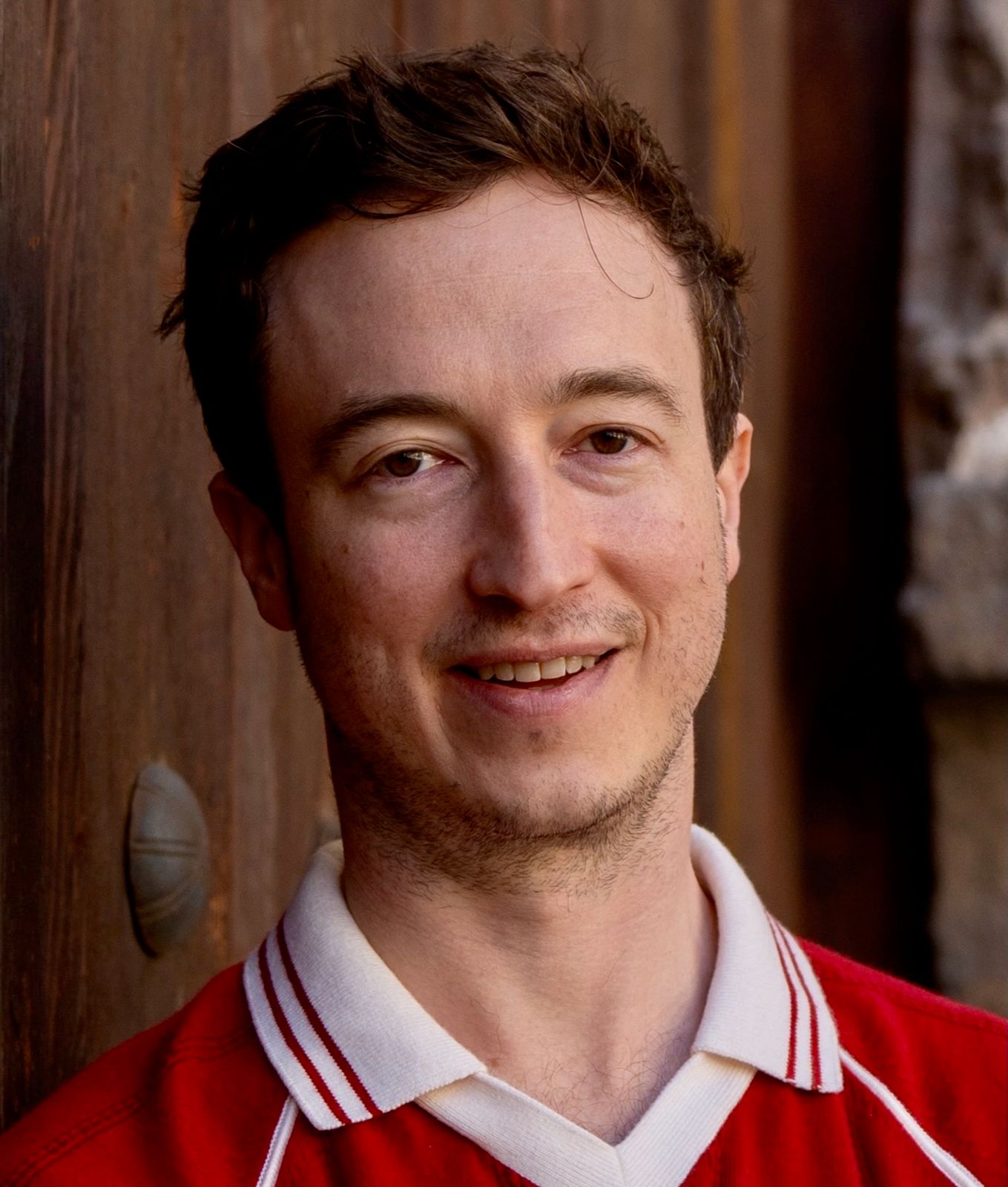 Liam smiling, in a red and white collared shirt, in front of a weathered wooden door.